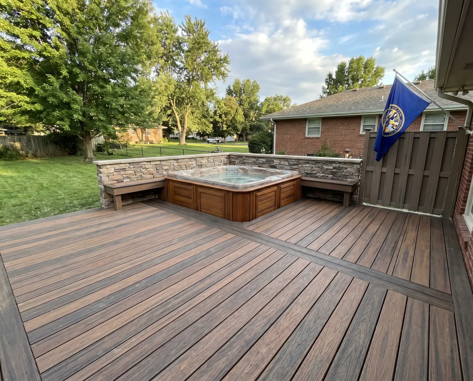 Backyard with a built‑in hot tub on a wooden deck, brick house to the right, and a blue flag near the fence amid green trees and a blue sky.