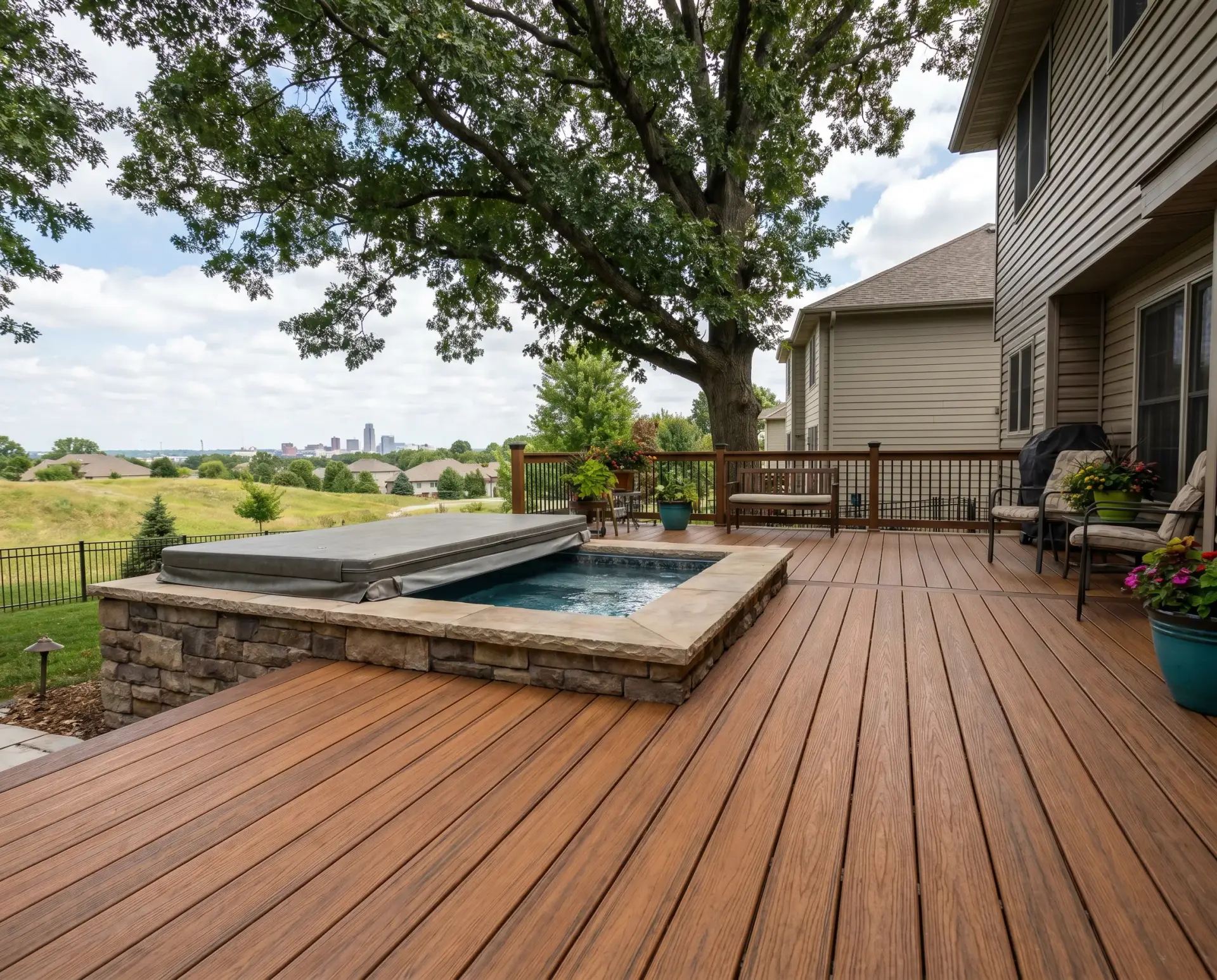 Wood deck with a built-in hot tub, railing, and a large tree providing shade; distant city skyline on the horizon.